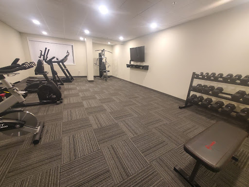 An empty fitness room with boring beige walls and grey flooring. Cardio machines line the left side of the room, an all-in-one weight machine is at the back next to a window, a large TV on the right hand side with freeweights and a bench in the foreground.